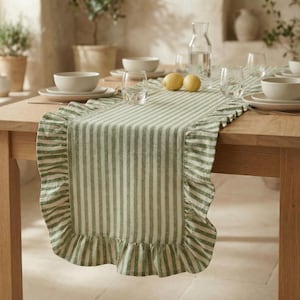 May include: A green and white striped table runner with a ruffled edge. The runner is placed on a wooden table, with white bowls, plates, and glasses visible. Two lemons are on the table.
