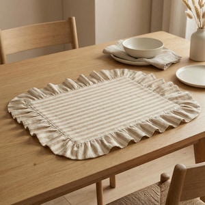 Op de afbeelding: Een rechthoekige beige en wit gestreepte placemat met een gerimpelde rand ligt op een lichte houten tafel. Een witte kom, bord en linnen servet liggen op de tafel. Een vaas met gedroogde bloemen staat ook op de tafel.