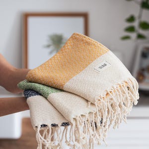May include: A stack of three woven cotton towels in shades of yellow, green, and white. Each towel features a textured diamond pattern and a fringed edge. The towels are labeled with the brand name "Wellsal".