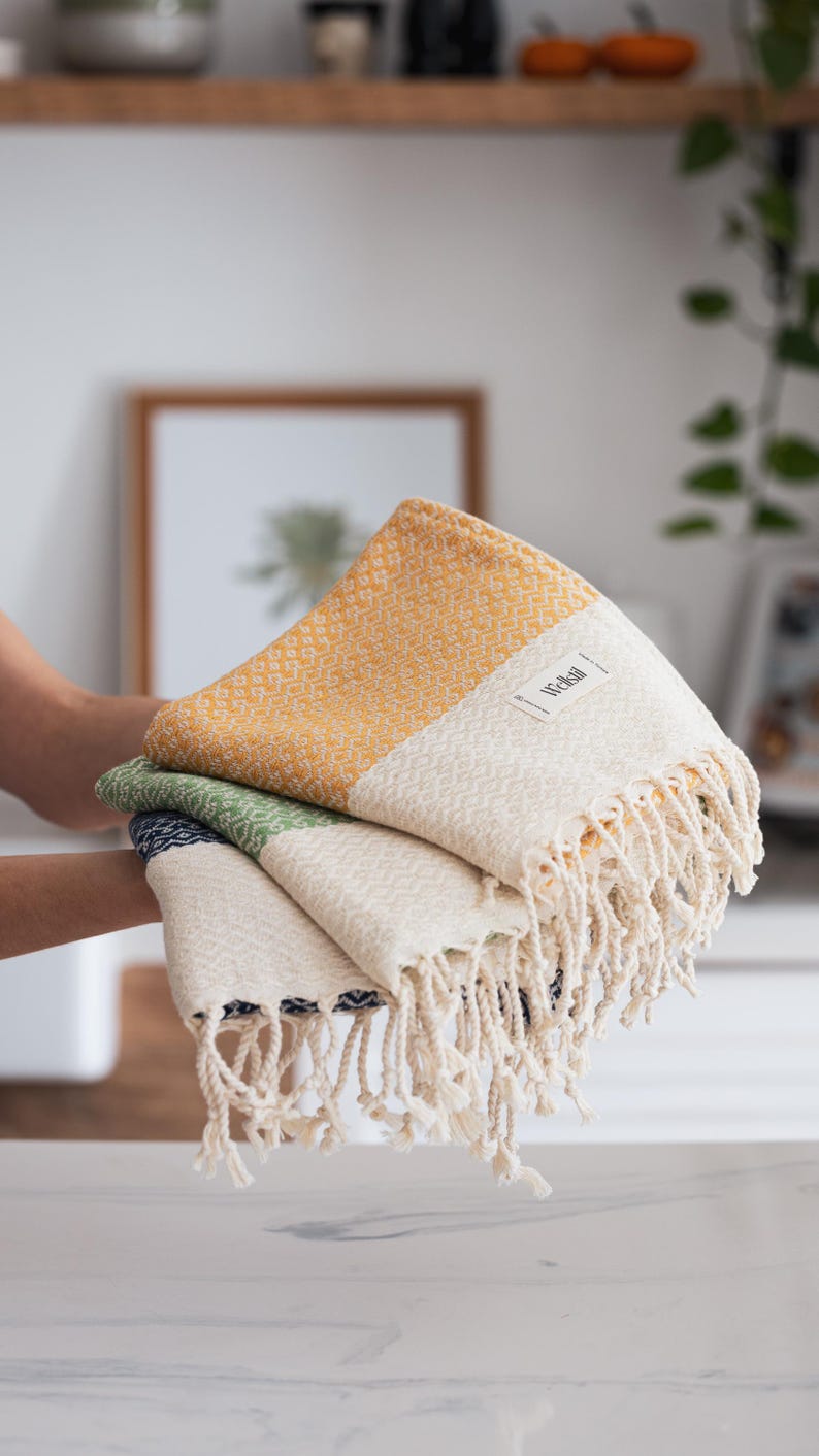 May include: A stack of three woven cotton towels in shades of yellow, green, and white. Each towel features a textured diamond pattern and a fringed edge. The towels are labeled with the brand name "Wellsal".