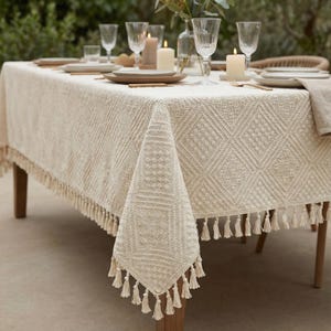 K&ouml;nnte beinhalten: Eine cremefarbene Tischdecke mit einem geometrischen Rautenmuster und Fransenbesatz. Der Tisch ist mit Tellern, Gl&auml;sern, Kerzen und Besteck gedeckt und schafft eine elegante Essszene. Die Tischdecke besteht aus einem strukturierten Stoff.