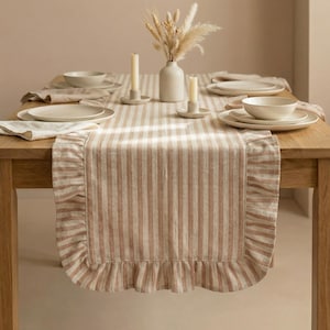 Puede incluir: Una mesa de comedor preparada para una comida, con un camino de mesa a rayas con borde con volantes. La mesa está puesta con platos blancos, cuencos, servilletas, velas y un jarrón con flores secas. La combinación de colores es neutra con beige y blanco.