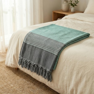 May include: A folded blanket with a teal, gray, and black color scheme, draped over a bed. The blanket has a textured weave and a fringe along the bottom edge. The bed has a white comforter.