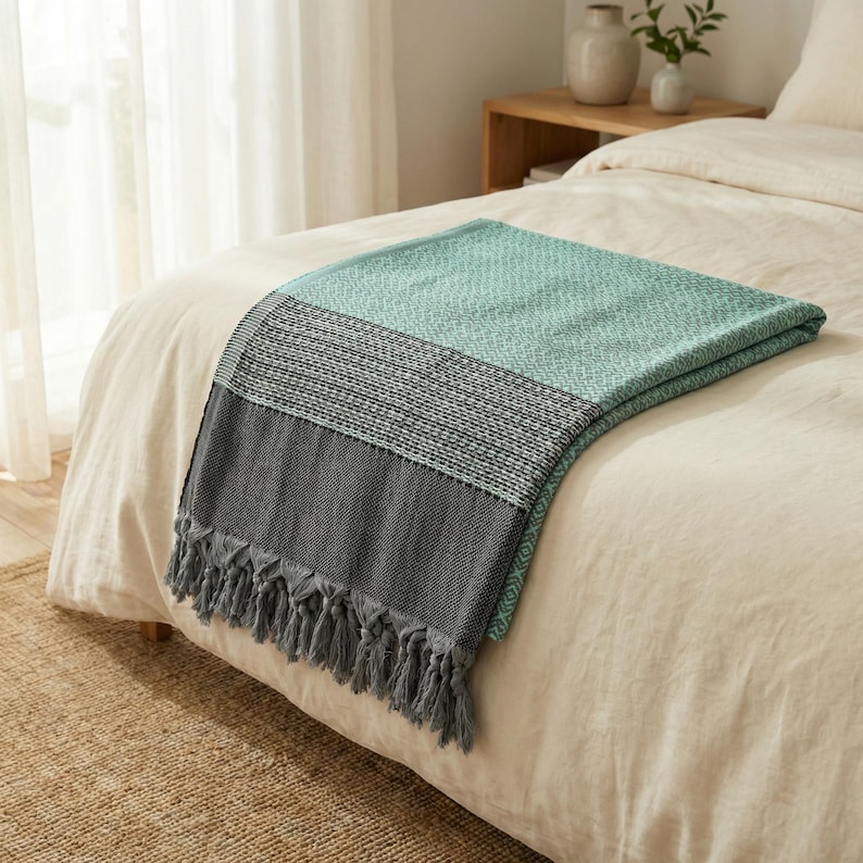 May include: A folded blanket with a teal, gray, and black color scheme, draped over a bed. The blanket has a textured weave and a fringe along the bottom edge. The bed has a white comforter.