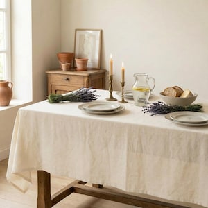 Könnte beinhalten: Ein Esstisch, der für eine Mahlzeit gedeckt ist. Der Tisch ist mit einer cremefarbenen Leinentischdecke bedeckt. Zwei brennende Kerzen in Messinghaltern, Teller, eine Wasserkaraffe, Brot und Lavendelzweige sind auf dem Tisch angeordnet.