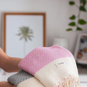 May include: Two folded Turkish towels in the image. One is pink and white, the other is gray and white. Both have a diamond pattern and fringe. The towels have a tag that says "Wellstil".
