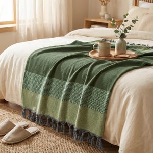 May include: A green throw blanket with a geometric pattern and gray fringe is draped over a bed. A wooden tray holds a mug, a coaster, and a small vase with greenery. Beige slippers rest on a woven rug.
