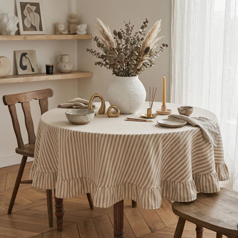 Op de afbeelding: Een ronde tafel met een gestreept tafelkleed met een gerimpelde rand. De tafel is gedekt met een witte vaas met gedroogde bloemen, een kaars en decoratieve voorwerpen. Een houten stoel staat aan de zijkant.