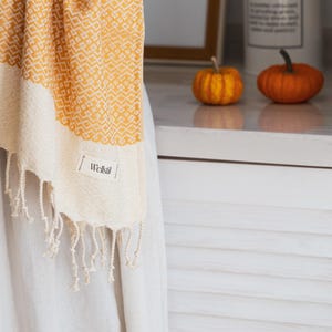 May include: A yellow and cream-colored woven towel with a geometric pattern and fringe. The towel has a label that reads "Wellstil". The towel is being held up in front of a white surface.