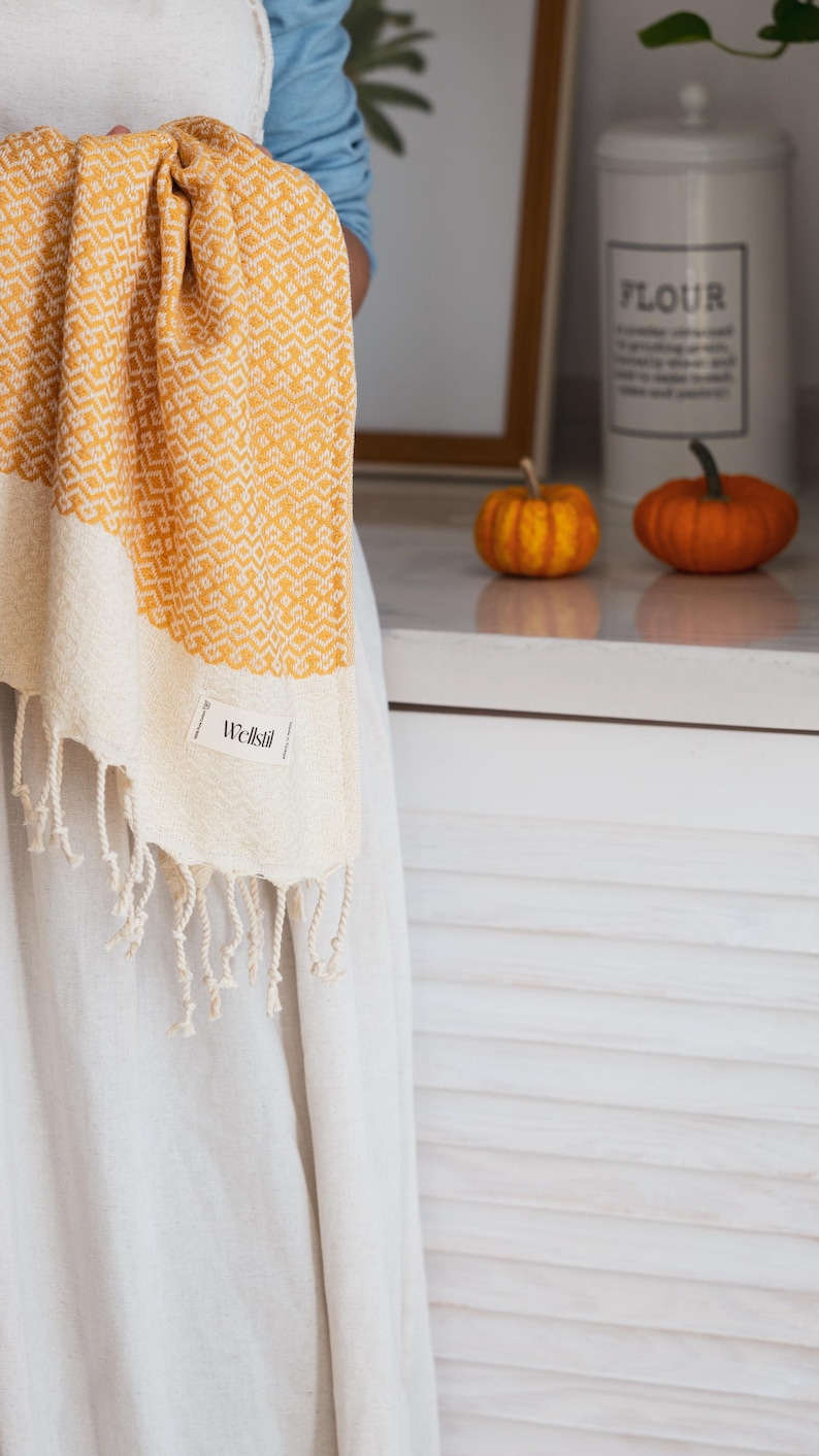 May include: A yellow and cream-colored woven towel with a geometric pattern and fringe. The towel has a label that reads "Wellstil". The towel is being held up in front of a white surface.