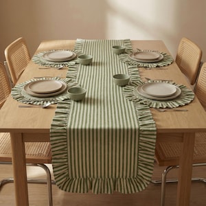 May include: A dining table set for a meal, featuring a green and white striped table runner with ruffled edges. Matching placemats and small bowls are arranged around the plates. Wicker chairs surround the wooden table, creating a cohesive dining aesthetic.