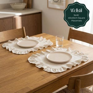 May include: Two off-white square placemats with ruffled edges and matching plates with a gold rim are set on a wooden table. A glass of water is also on the table. The placemats are labeled "Wellstil" and are 16x16 inches (40x40cm).