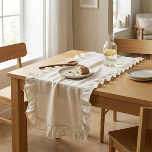 May include: A beige linen table runner with a ruffled edge is laid on a wooden table. A plate with bread, a wooden spoon, and a glass water pitcher with lemon slices are on the table. Two wooden chairs are visible.
