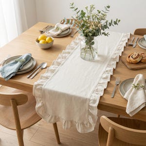 May include: A wooden dining table set for a meal, featuring a cream-coloured ruffled table runner. The table is set with plates, silverware, napkins, and a vase of flowers. A bowl of lemons and bread rolls are also present.