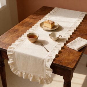 May include: A cream-colored linen table runner with ruffled edges, set on a rustic wooden table. A small plate with a slice of cake, a teacup, a small bowl, a fork, and a folded napkin are arranged on the runner, creating a cozy dining scene.