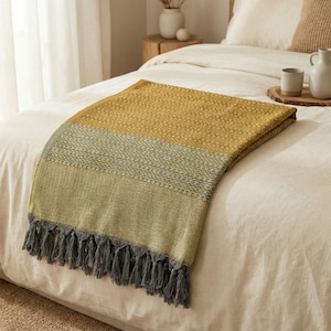 May include: A folded mustard yellow and green throw blanket with a blue stripe and gray fringe. The blanket is made of a woven fabric and is displayed on a white bed.