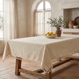K&ouml;nnte beinhalten: Cremefarbene rechteckige Tischdecke mit einem strukturierten Rautenmuster und Fransenbesatz. Die Tischdecke ist &uuml;ber einem rustikalen Holztisch drapiert. Eine Sch&uuml;ssel mit Zitronen, eine Vase mit Olivenzweigen und andere dekorative Gegenst&auml;nde befinden sich auf dem Tisch.