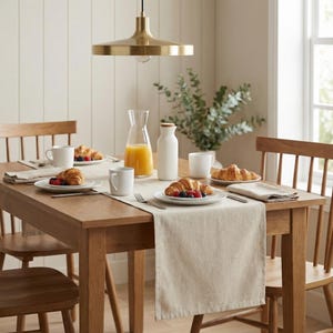 May include: A wooden dining table set for breakfast with croissants, berries, and juice. The table is set with white plates, mugs, and a beige runner. A gold-colored light fixture hangs above the table, and wooden chairs surround it.
