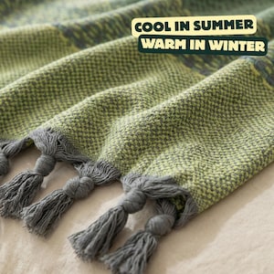 May include: Close-up of a woven blanket in shades of green and gray, with decorative tassels at the edge. The blanket has a textured appearance and the text "COOL IN SUMMER WARM IN WINTER".