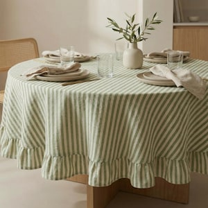 Può includere: Un tavolo rotondo apparecchiato per la cena, con una tovaglia a righe verdi e bianche con bordo a balze. Il tavolo è apparecchiato con piatti, tovaglioli e bicchieri. Un vaso con rami d'ulivo aggiunge un tocco naturale.