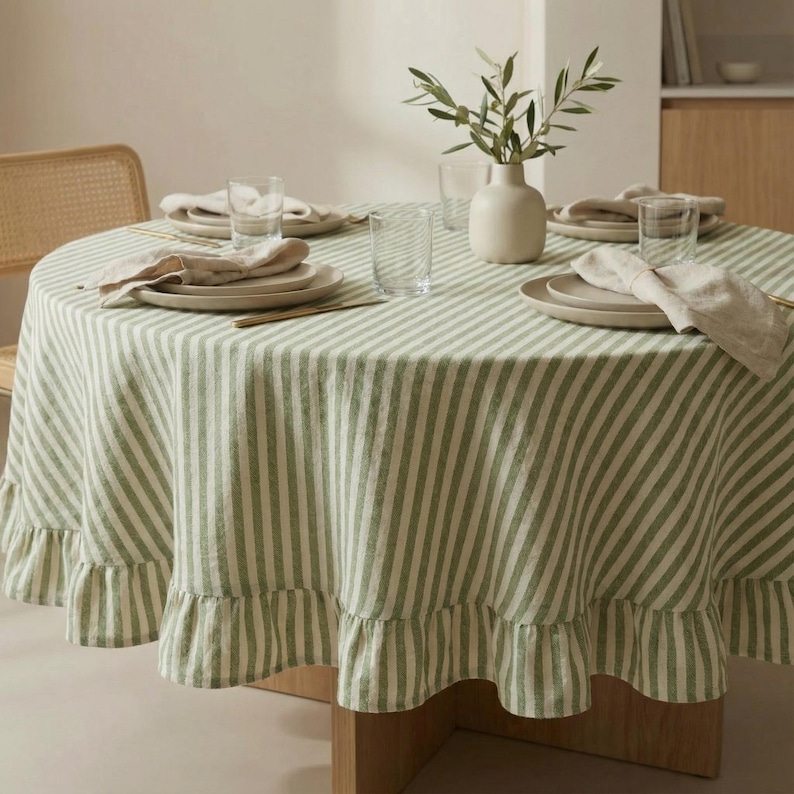 May include: A round table set for dining, featuring a green and white striped tablecloth with a ruffled edge. The table is set with plates, napkins, and glasses. A vase with olive branches adds a natural touch to the setting.
