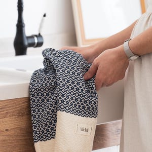 May include: A navy blue and cream-colored woven towel with a geometric pattern is held near a white sink. The towel has a cream-colored fringe and a label that reads "Wellsi". A black faucet is visible in the background.