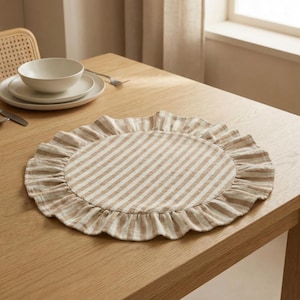 Op de afbeelding: Een ronde placemat van stof met een beige en wit verticaal gestreept patroon en een geplooide, geruffelde rand. De placemat is gemaakt van een getextureerd textielmateriaal en ligt plat op een houten oppervlak, gecentreerd in het beeld.