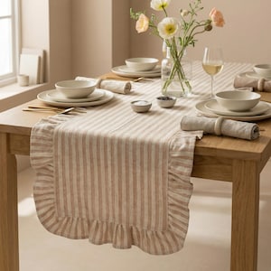 Könnte beinhalten: Ein Holztisch, gedeckt für eine Mahlzeit, mit einem gestreiften Tischläufer mit Rüschenkante. Der Tisch ist mit weißen Tellern, Schalen und Servietten sowie goldenem Besteck und einer Blumenvase gedeckt.