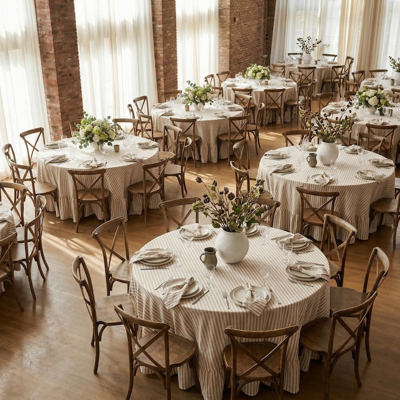 Op de afbeelding: Een bovenaanzicht van een grote evenementenruimte met meerdere ronde tafels die zijn opgemaakt voor een bijeenkomst. Elke tafel is bedekt met een gestreept tafelkleed en omringd door houten stoelen. Bloemen en vazen sieren de tafels.