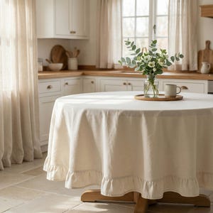May include: A round, cream-colored tablecloth with a ruffled edge covers a wooden table. A clear glass vase with greenery and a mug sit on a wooden tray in the center. The kitchen has white cabinets and a window.