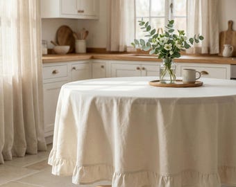 May include: A round, cream-colored tablecloth with a ruffled edge covers a wooden table. A clear glass vase with greenery and a mug sit on a wooden tray in the center. The kitchen has white cabinets and a window.