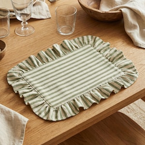 May include: A rectangular placemat with a green and white striped pattern and a ruffled edge. The placemat is made of fabric and is placed on a wooden table. The placemat is designed for dining and adds a decorative touch to the table setting.