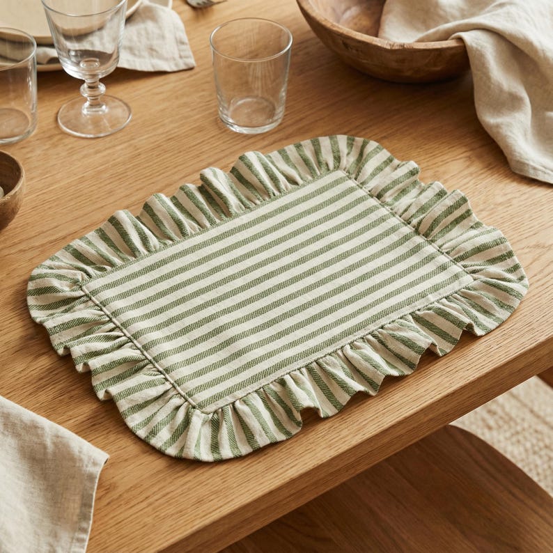 May include: A rectangular placemat with a green and white striped pattern and a ruffled edge. The placemat is made of fabric and is placed on a wooden table. The placemat is designed for dining and adds a decorative touch to the table setting.