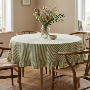 May include: A round dining table with a green and white striped tablecloth. The tablecloth has a ruffled edge. The table is surrounded by wooden chairs. A vase of flowers sits on the table, along with a small bowl.