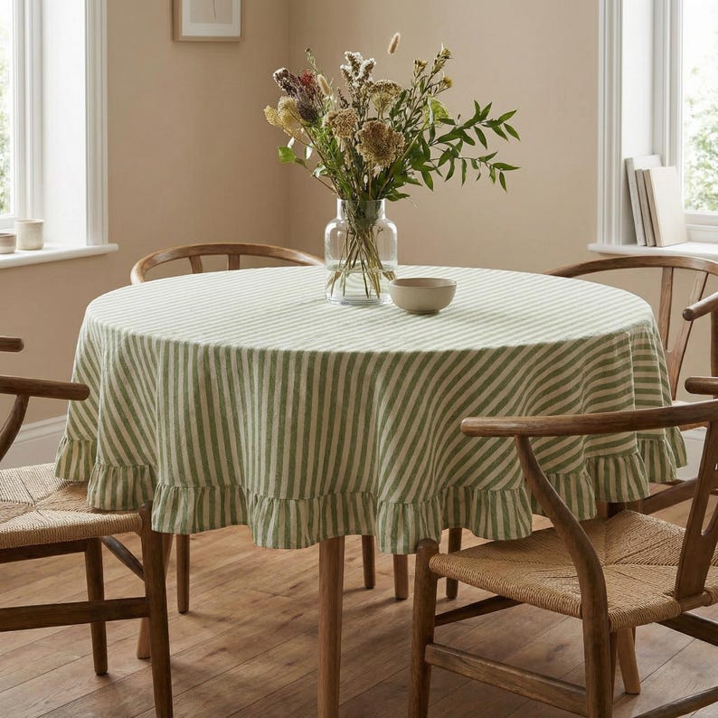 May include: A round dining table with a green and white striped tablecloth. The tablecloth has a ruffled edge. The table is surrounded by wooden chairs. A vase of flowers sits on the table, along with a small bowl.