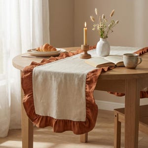 Pode incluir: Caminho de mesa de linho bege com borda de babados terracota. O caminho de mesa está colocado sobre uma mesa redonda de madeira, junto com uma vela acesa, um vaso de flores secas e uma caneca. Um croissant está em um prato.