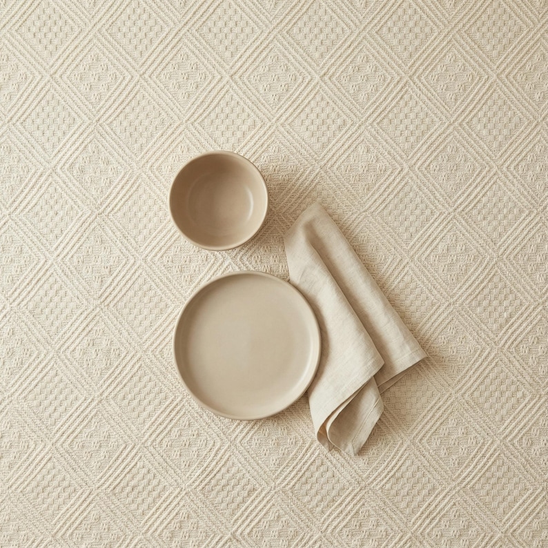 K&ouml;nnte beinhalten: Eine beige Keramiksch&uuml;ssel und ein Teller mit einer gefalteten Leinen-Serviette auf einer cremefarbenen Tischdecke mit Rautenmuster. Das Geschirr ist f&uuml;r eine schlichte, neutral gehaltene Tischdekoration arrangiert.