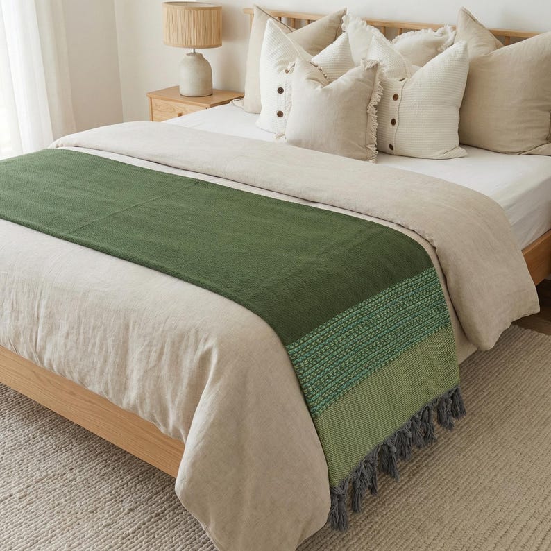 May include: A green bed runner with a textured design and gray tassels. The runner is placed on a bed with beige bedding and pillows. The bed frame is made of light-colored wood.