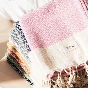 May include: A stack of colorful woven Turkish towels with fringe. The top towel is pink and white, with a diamond pattern. Other towels are in shades of blue, green, yellow, and orange. The towels are labeled "Wellstil".