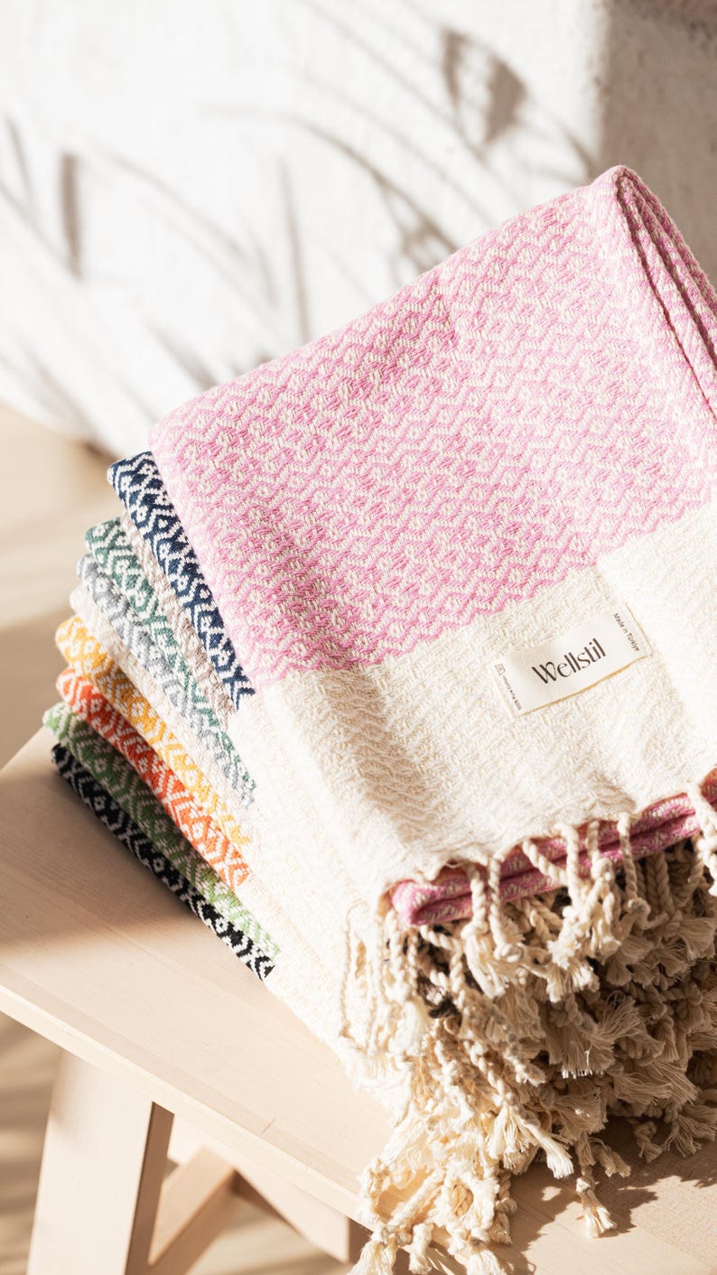 May include: A stack of colorful woven Turkish towels with fringe. The top towel is pink and white, with a diamond pattern. Other towels are in shades of blue, green, yellow, and orange. The towels are labeled "Wellstil".