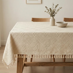 May include: Cream-coloured rectangular tablecloth with a diamond pattern and fringe trim. The tablecloth is draped over a wooden table, with a vase of greenery and a bowl on top. The overall aesthetic is neutral and rustic.