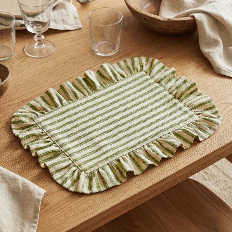 May include: A rectangular placemat with a green and white striped pattern and a ruffled edge. The placemat is made of fabric and is placed on a wooden table, alongside glassware and a wooden bowl. The overall theme is dining and home decor.