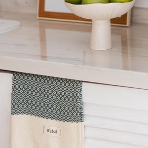 May include: A green and white patterned towel with fringe hangs from a white cabinet. The towel has a geometric design in green and a cream-colored bottom section. A bowl of green fruit sits on the countertop above.