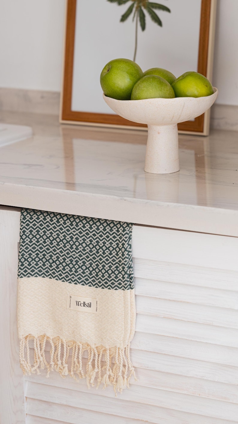 May include: A green and white patterned towel with fringe hangs from a white cabinet. The towel has a geometric design in green and a cream-colored bottom section. A bowl of green fruit sits on the countertop above.