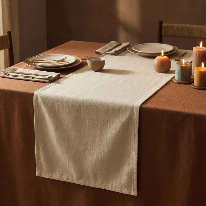 May include: A dining table set for a meal, featuring a beige table runner, plates, napkins, and lit candles. The table is covered with a brown tablecloth. The scene evokes a warm, inviting atmosphere, perfect for a dinner party or special occasion.