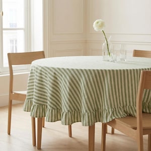 May include: A round tablecloth with green and white stripes and a ruffled edge. The table is set with wooden chairs, a vase with a white flower, and two clear glasses. The room has a window.