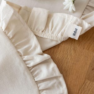 May include: Close-up of a cream-colored apron with ruffled edges. The apron has a tag that reads "Wellstil". A white daisy and a plate of cookies are in the background. The apron is made of a soft, natural fabric.