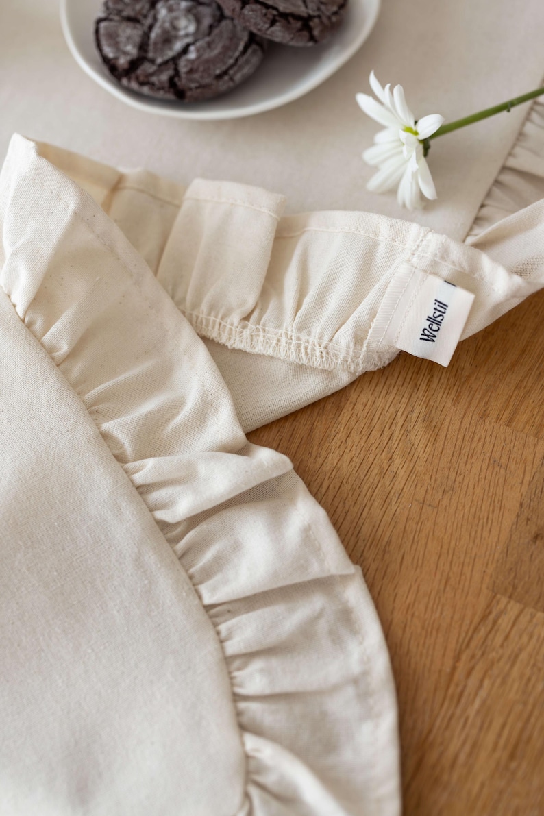 May include: Close-up of a cream-colored apron with ruffled edges. The apron has a tag that reads "Wellstil". A white daisy and a plate of cookies are in the background. The apron is made of a soft, natural fabric.