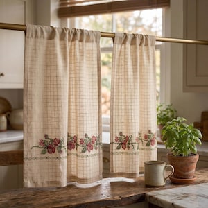 May include: Two beige kitchen curtains with a grid pattern and floral embroidery along the bottom. The curtains are hanging on a gold rod in front of a window. A mug and potted plant sit on the wooden counter.