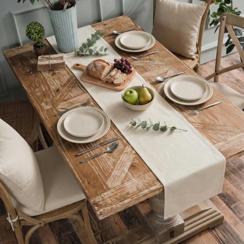 May include: A rustic wooden dining table set for a meal. The table is adorned with a cream-colored runner, plates, silverware, bread, fruit, and decorative greenery. The table is surrounded by chairs with cream-colored cushions.